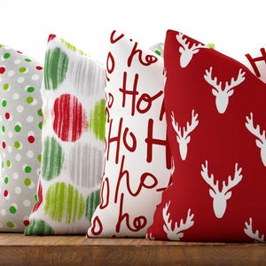 May include: Four decorative Christmas pillows with festive patterns. Designs include polka dots, brushstrokes, "Ho Ho Ho" text, and white reindeer silhouettes on a red background. The pillows are arranged on a wooden surface.