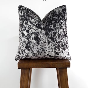 May include: A black and white cowhide pillow with a salt and pepper pattern. The pillow is sitting on a wooden stool.