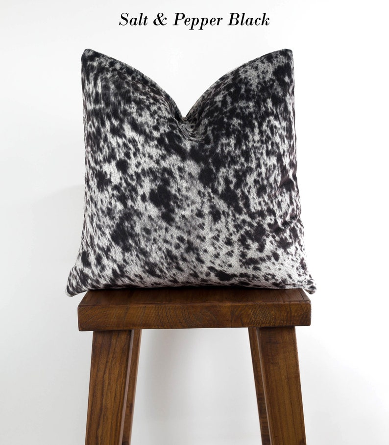 May include: A black and white cowhide pillow with a salt and pepper pattern. The pillow is sitting on a wooden stool.
