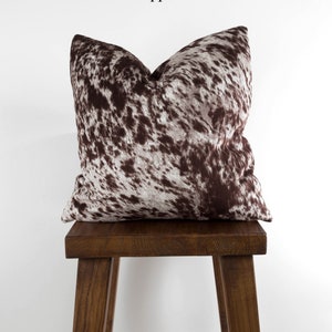 May include: A brown and white cowhide patterned throw pillow on a wooden stool. The pillow has a brown and white pattern that resembles cowhide. The stool is made of dark wood.