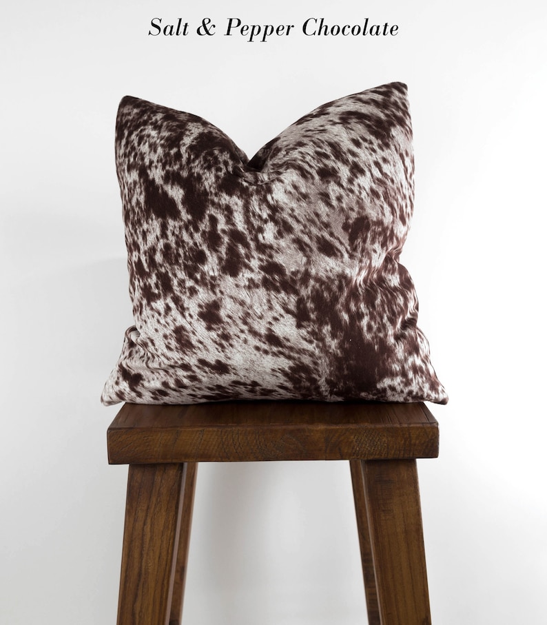 May include: A brown and white cowhide patterned throw pillow on a wooden stool. The pillow has a brown and white pattern that resembles cowhide. The stool is made of dark wood.