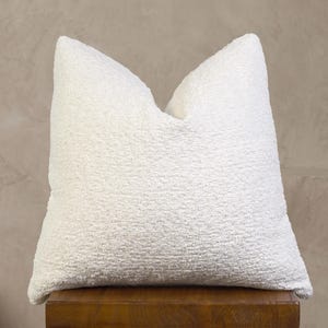 May include: A square throw pillow featuring a textured, off-white bouclé fabric cover. The surface of the pillow has a distinct nubby, looped appearance, creating a tactile and dimensional look across the entire front face of the cushion.