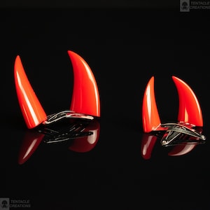 Puede incluir: Dos pares de pinzas para el pelo con forma de cuernos de diablo rojos. El par más grande está en el fondo y el par más pequeño está en primer plano. Ambos pares están sobre una superficie negra.