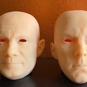 May include: Two realistic, pale-skinned human head masks with red eye sockets. The masks have detailed facial features, including noses, mouths, and ears. The masks are displayed against a brown background.