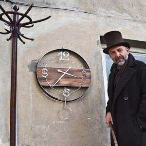 Peut inclure: Un homme portant un manteau noir et un chapeau haut de forme se tient devant un mur avec une horloge. L'horloge a un cadran en bois avec des accents métalliques et des chiffres romains. L'homme tient une canne.