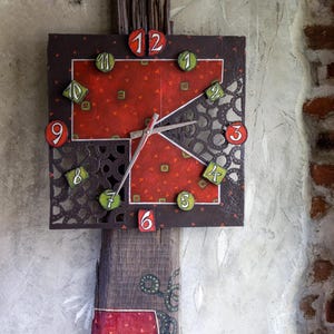 May include: A handcrafted wall clock with a unique design. The clock features a square face with a red patterned background and black metal cutouts. The numbers are in green and red. The clock is mounted on a wooden base.