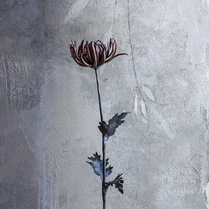 Könnte beinhalten: Eine Metallsculptur einer Blume mit braunen Blütenblättern und grauen Blättern auf einem weißen Steinsockel.