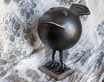 Op de afbeelding: Een zwart metalen vogelsculptuur met een rond lichaam en lange poten. De vogel staat op een kleine metalen voet.