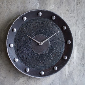 May include: A round, black clock with silver accents. The clock face has a concentric circle design and silver hands. The clock is mounted on a gray wall.