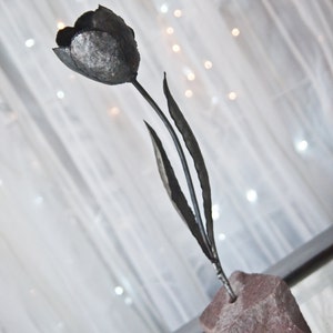 Op de afbeelding: Een metalen sculptuur van een tulp met een donkergrijze afwerking. Het beeld staat op een kleine, ruwe stenen basis.