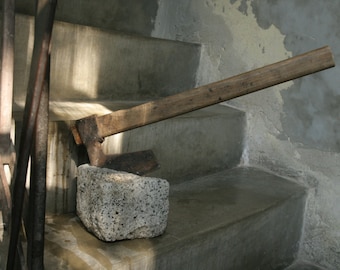 May include: A wooden-handled axe with a metal blade rests on a concrete staircase. The axe is resting on a grey stone block.