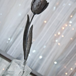 Op de afbeelding: Een metalen sculptuur van een enkele tulp in een heldere glazen vaas. De tulp is gemaakt van donkergrijs metaal en heeft een lange steel en bladeren.