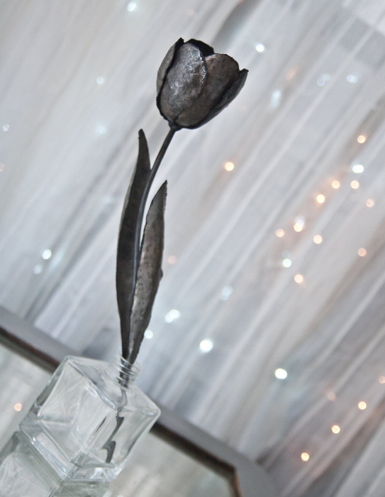 Op de afbeelding: Een metalen sculptuur van een enkele tulp in een heldere glazen vaas. De tulp is gemaakt van donkergrijs metaal en heeft een lange steel en bladeren.