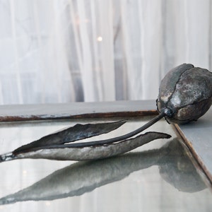 Op de afbeelding: Een metalen sculptuur van een tulp met een grijze en bruine afwerking. Het beeldje ligt op een glazen oppervlak.