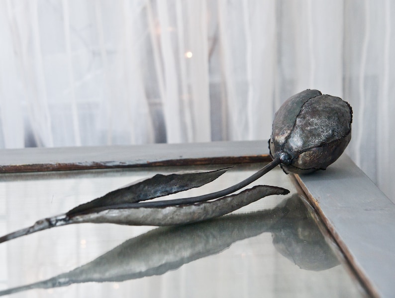 Op de afbeelding: Een metalen sculptuur van een tulp met een grijze en bruine afwerking. Het beeldje ligt op een glazen oppervlak.
