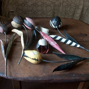 May include: A collection of handmade metal tulips with painted details. The tulips are in various colors, including red, white, black, yellow, and pink. The stems are black metal and have leaves.