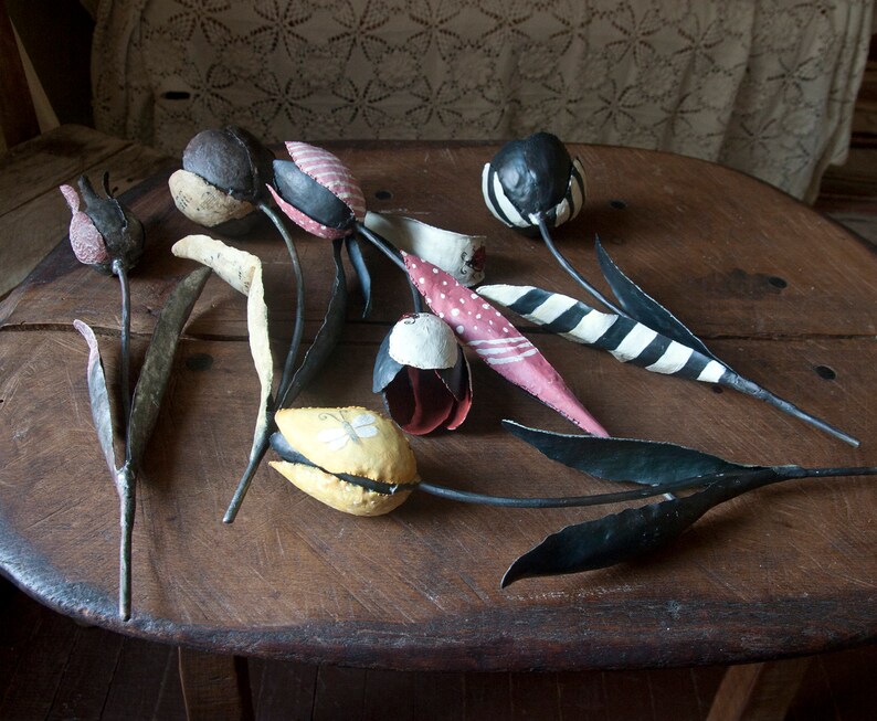 May include: A collection of handmade metal tulips with painted details. The tulips are in various colors, including red, white, black, yellow, and pink. The stems are black metal and have leaves.