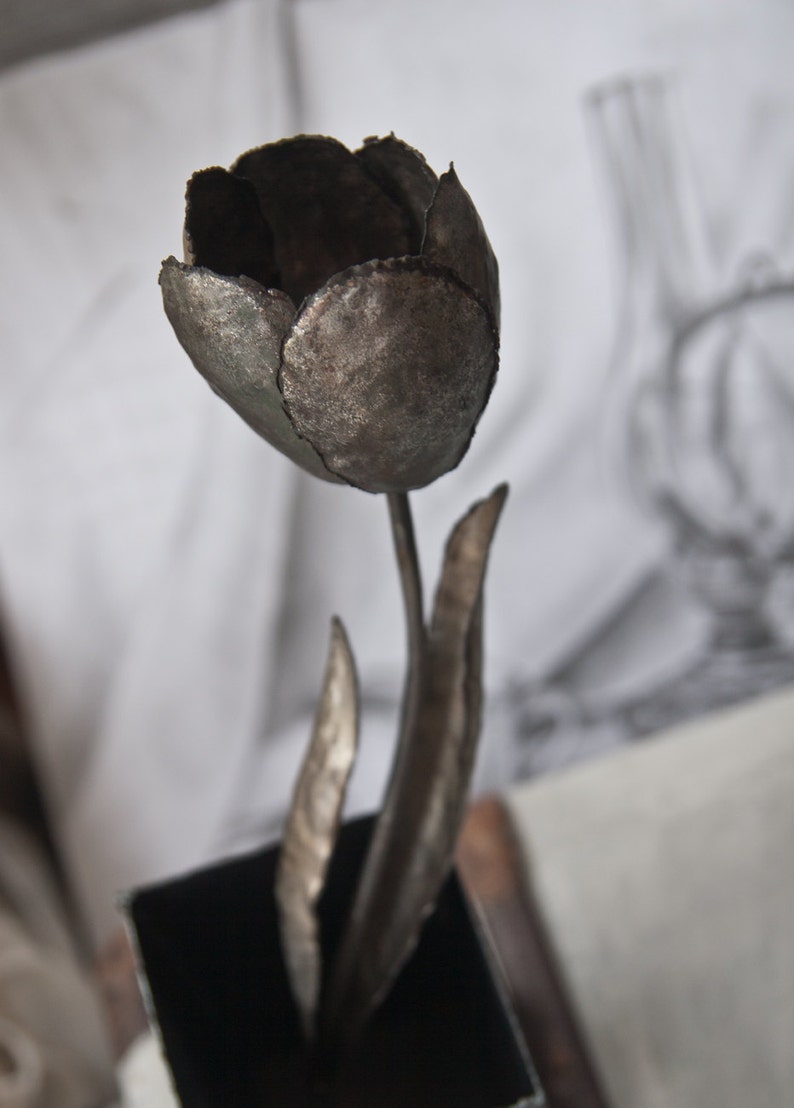Op de afbeelding: Een metalen sculptuur van een tulp met een donkergrijze afwerking. De tulp staat in een zwarte vierkante pot.
