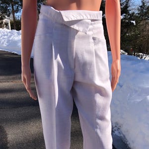 Op de afbeelding: Witte linnen broek met een unieke wikkelband. De broek heeft een lichte, neutrale kleur en een getailleerde pasvorm. Het model draagt een roze top. De broek wordt op een zonnige dag buiten op een mannequin gepresenteerd.