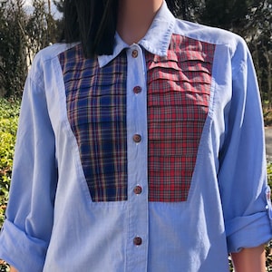 May include: A light blue long-sleeve button-down shirt featuring a contrasting front panel with vertical pleats. The panel is divided into two sections, one with a blue and yellow plaid pattern and the other with a red and green plaid pattern.