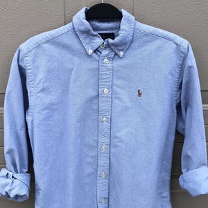 May include: Light blue button-down shirt with long sleeves, rolled up at the cuffs. The shirt features a button-down collar and a small embroidered logo on the chest. The shirt is hanging on a black hanger.