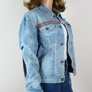 May include: Light blue denim jacket with a classic collar, button closure, and pockets. Features a plaid accent on the chest pocket and purple stitching details. The jacket is paired with dark blue jeans.