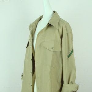 May include: A beige long-sleeved shirt with a button-down collar and a green patch on the sleeve.