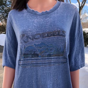 May include: Blue short-sleeved t-shirt with the word "KNOEBELS" printed across the top. Below the word is a graphic of a mountain range and a roller coaster. The shirt is made of a soft, worn material.