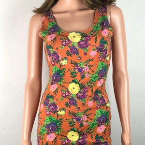 May include: Orange tank dress with a floral pattern. The dress features a scoop neckline and is covered in a repeating pattern of purple, pink, and yellow flowers with green leaves. The dress is form-fitting and appears to be made of a stretchy fabric.