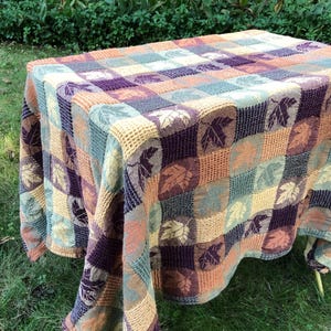 May include: A rectangular tablecloth featuring a checkered pattern of squares in shades of purple, orange, teal, and cream. Each square displays a detailed leaf motif. The fabric has a textured, waffle-like weave throughout the entire surface.