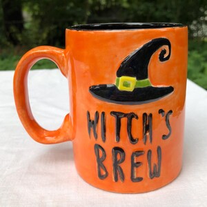 May include: A bright orange ceramic mug with a black interior and a handle. The front features a raised black witch hat with a green band and yellow buckle, and the black embossed text reads WITCH'S BREW in a stylized, hand-lettered font.