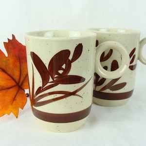 May include: Two brown and white ceramic mugs with a brown floral design. The mugs have a speckled glaze and a brown band around the top.