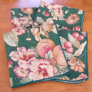 May include: Four folded green cloth napkins with a floral and fruit pattern. The design includes pink and peach flowers, peaches, cherries, and strawberries. The napkins have a teal border and are arranged on a wooden surface.