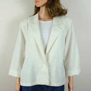 May include: A light beige blazer with three-quarter sleeves and a single-button closure. The blazer has a notched collar and is made of a textured fabric. The sleeves are cuffed at the wrists. The blazer is worn over a white top.