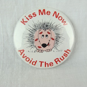 May include: A white button with a red border and the text "Kiss Me Now Avoid The Rush". A cartoon pink hedgehog with red lips and black eyes is in the center of the button.