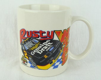 May include: White ceramic mug featuring a black and yellow race car illustration. The car has the words "Miller Genuine Draft" on the side. The text "Rusty Wallace" is in red and white on the mug.
