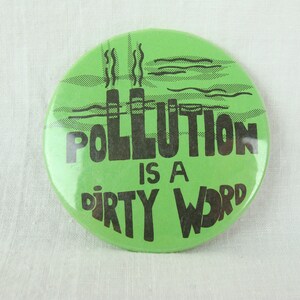 May include: Green button with black text that reads "Pollution is a dirty word". The button features a graphic of two smokestacks with smoke coming out of them.