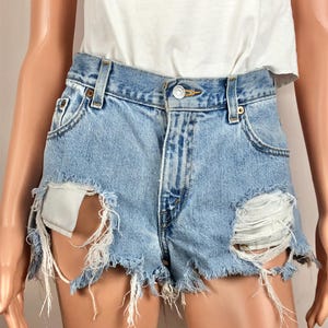 May include: Light blue denim shorts with distressed detailing, featuring large rips and frayed edges. The shorts have a classic five-pocket design and a button closure. The shorts are part of a casual clothing collection.