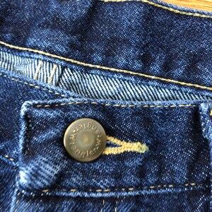 Op de afbeelding: Close-up van een blauwe denim jeansknoop met het woord "Wrangler" in goud gegraveerd.