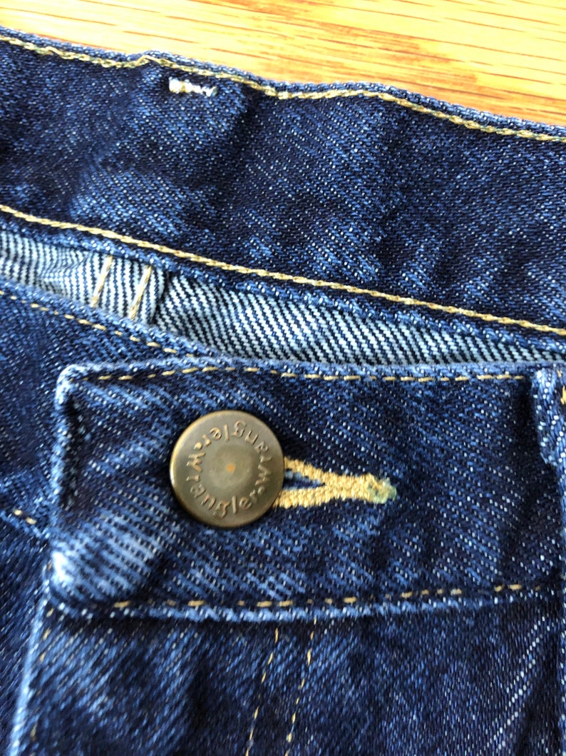Op de afbeelding: Close-up van een blauwe denim jeansknoop met het woord "Wrangler" in goud gegraveerd.