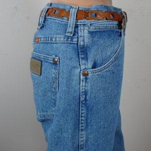 May include: Light blue denim jeans with a brown leather belt. The jeans have a classic five-pocket design, a brown leather patch on the back pocket, and copper rivets. The belt features a decorative cut-out pattern.