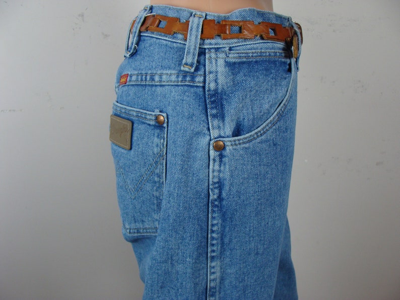 May include: Light blue denim jeans with a brown leather belt. The jeans have a classic five-pocket design, a brown leather patch on the back pocket, and copper rivets. The belt features a decorative cut-out pattern.