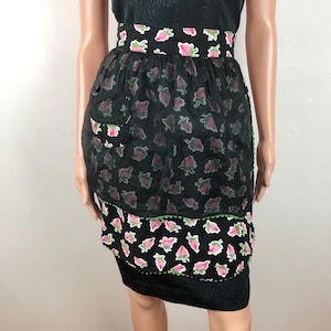 Op de afbeelding: Een zwarte schort met roze bloemenprint, een transparante overlay en een groene bies. De schort heeft een grote zak aan de voorkant.