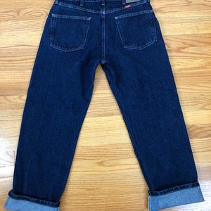 Op de afbeelding: Een donkerblauwe spijkerbroek met een Wrangler-label op de achterzak. De broek heeft een gezoomde zoom.