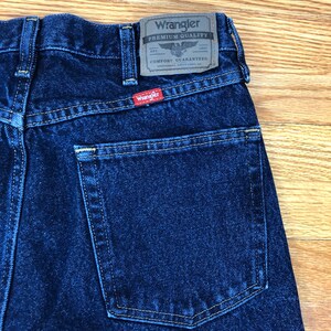 Op de afbeelding: Een donkerblauwe spijkerbroek met een achterzak en een Wrangler-label met de tekst "Premium Quality" met een adelaarlogo en "Comfort Guaranteed".