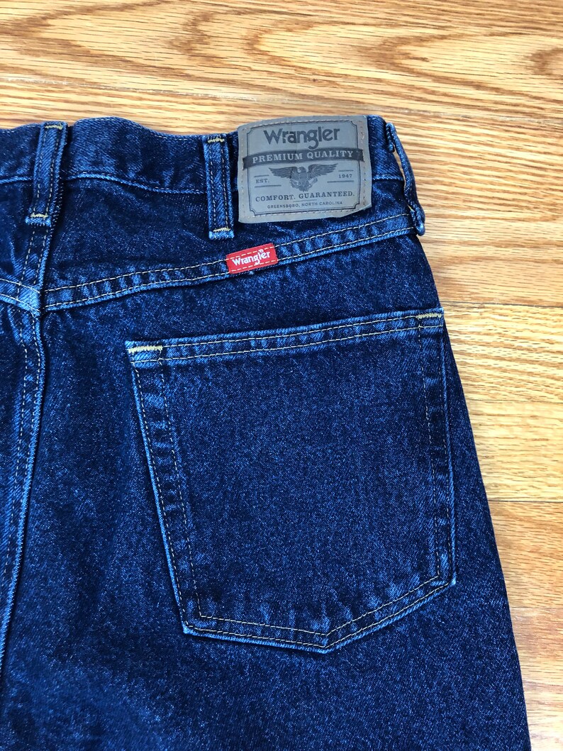 Op de afbeelding: Een donkerblauwe spijkerbroek met een achterzak en een Wrangler-label met de tekst "Premium Quality" met een adelaarlogo en "Comfort Guaranteed".
