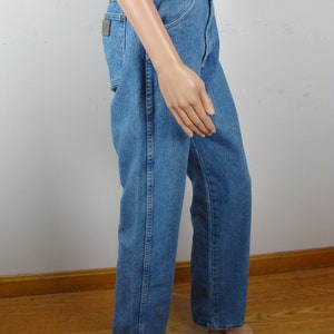 May include: Light blue denim jeans with a classic straight leg fit. The jeans feature a traditional five-pocket design, a button and zipper closure, and a small brown patch on the back pocket. The jeans are displayed on a mannequin.