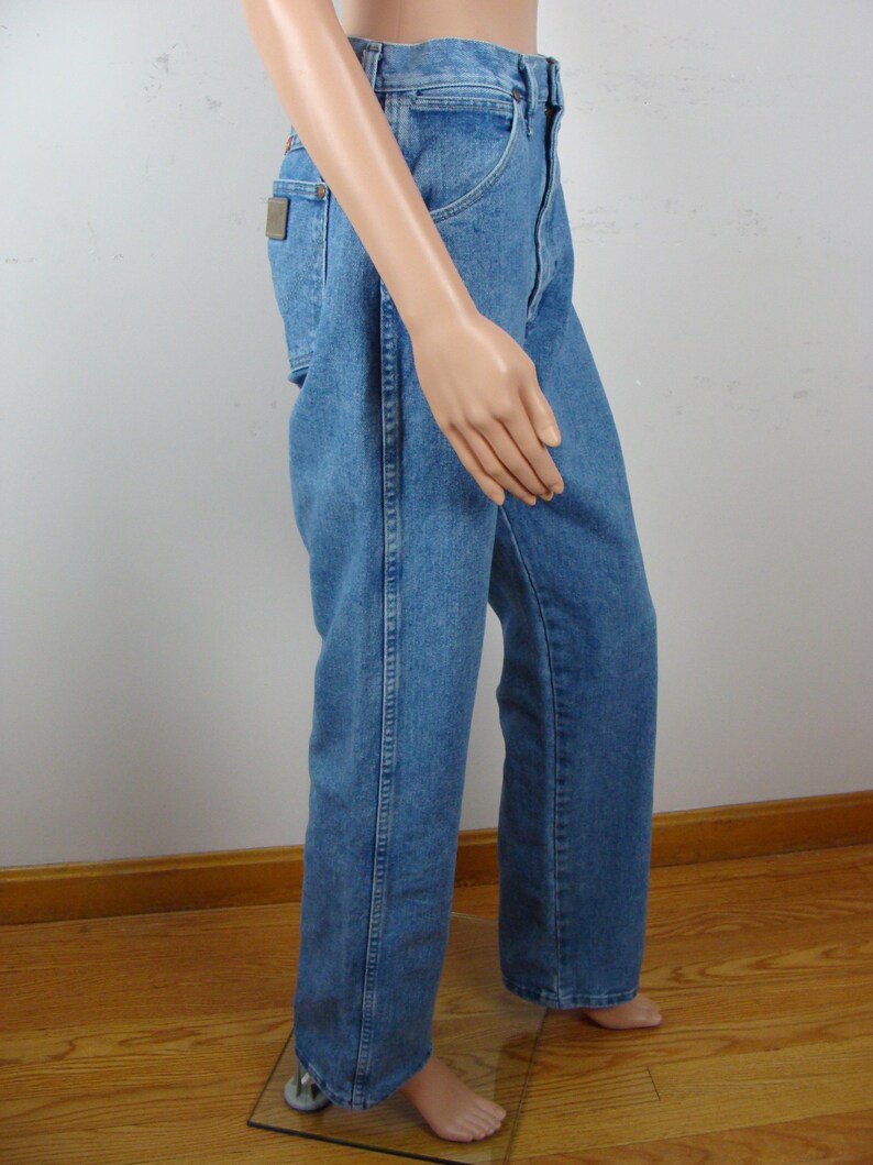 May include: Light blue denim jeans with a classic straight leg fit. The jeans feature a traditional five-pocket design, a button and zipper closure, and a small brown patch on the back pocket. The jeans are displayed on a mannequin.