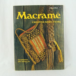 May include: A vintage book titled "Macrame: Creative Knot-Tying" with a brown wooden background. The cover features a macrame bag with a brown leather strap and tassels.