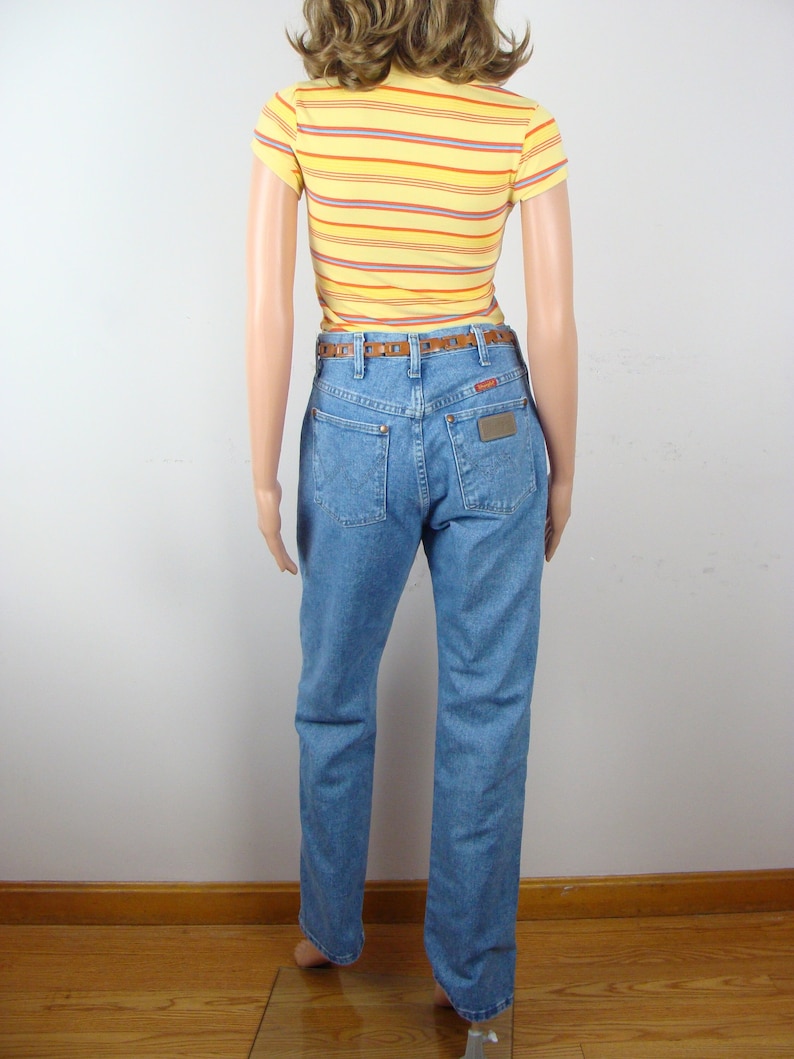 May include: A mannequin wearing a yellow striped t-shirt, light blue jeans, and a brown belt. The jeans have a Wrangler label on the back pocket. The t-shirt has horizontal stripes in orange, purple, and blue.